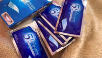 Multiple 5D White Teeth Whitening Strips packets and boxes scattered on a textured beige surface - mint flavour, enamel safe with professional effects
