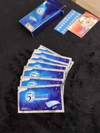 5D White Teeth Whitening Strips packets fanned out on a dark surface with product box and teeth shade guide card in the background