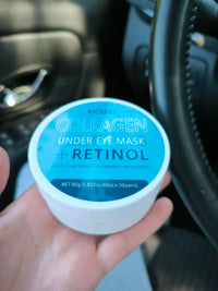 A hand holding a white round jar of XIOIMY Collagen Under Eye Mask + Retinol, with a blue label, in front of a dark car interior background.