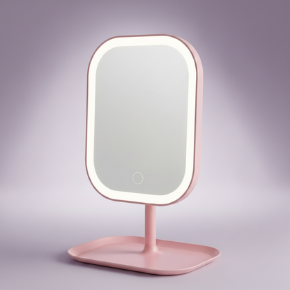 A pink vanity mirror with a rounded rectangular face, illuminated white LED border, and matching pink stand with a tray base, shown on a soft lavender studio background.