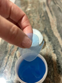 Fingers holding a translucent blue under-eye patch above an open jar on a patterned surface