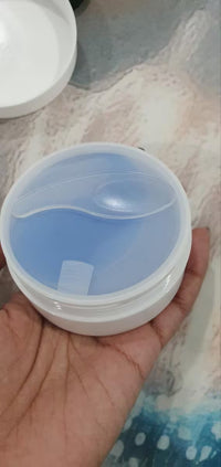 A hand holding a small round white plastic container with a translucent light blue lid or insert, photographed over a patterned surface.