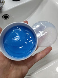 A hand holding an open white plastic jar filled with bright blue gel patches, with a clear lid resting beside it on a white sink surface.
