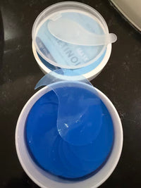 Translucent under-eye patch draped over an open jar of blue patches, with the lid and spatula behind it.