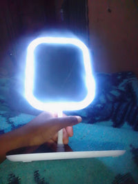 A handheld LED vanity mirror with a glowing white light frame and a white stand, held by a hand against a dark, dimly lit background.