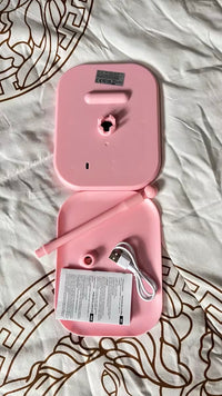 Pink electronic device shown open, revealing accessories including a pen-like wand, USB charging cable, small cap, and printed manual, placed on a white patterned fabric surface.