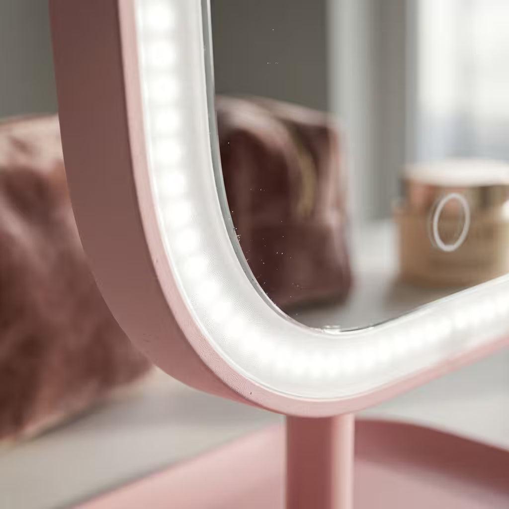 A close-up of a pink vanity mirror with a built-in LED light ring, showing a softly blurred makeup bag and cosmetic jar in the reflection.