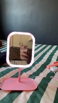 A pink vanity mirror with a built-in LED light ring and a matching pink stand, shown on a green and white striped bedspread with a mirror reflection of a person taking the photo.