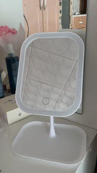 A white LED vanity mirror with a square frame and adjustable stand, shown on a white tabletop with a mirror and room background.
