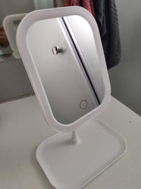 A white LED vanity mirror with a rounded rectangular frame and a matching square base, shown on a light-colored tabletop with a mirror reflection and a towel in the background.