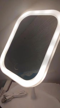 A white LED vanity mirror with a rounded rectangular shape, illuminated from the bright white light frame, shown on a white surface with its power cable visible.