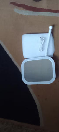 A white electronic device with a square mirror-like glass panel and a small white stylus, photographed on a patterned brown surface.