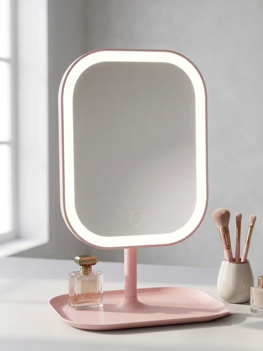 A pink LED vanity mirror with a built-in light and a matching tray base, styled on a white surface with cosmetic items and a perfume bottle in a bright room.